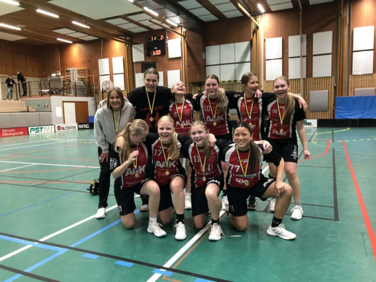 Ett ungdomslag i innebandy poserar tillsammans på en idrottshall efter en match. Flera spelare bär medaljer och matchtröjor, och några sitter på huk längst fram medan resten står bakom. I bakgrunden syns innebandyplanen och resultattavlan.