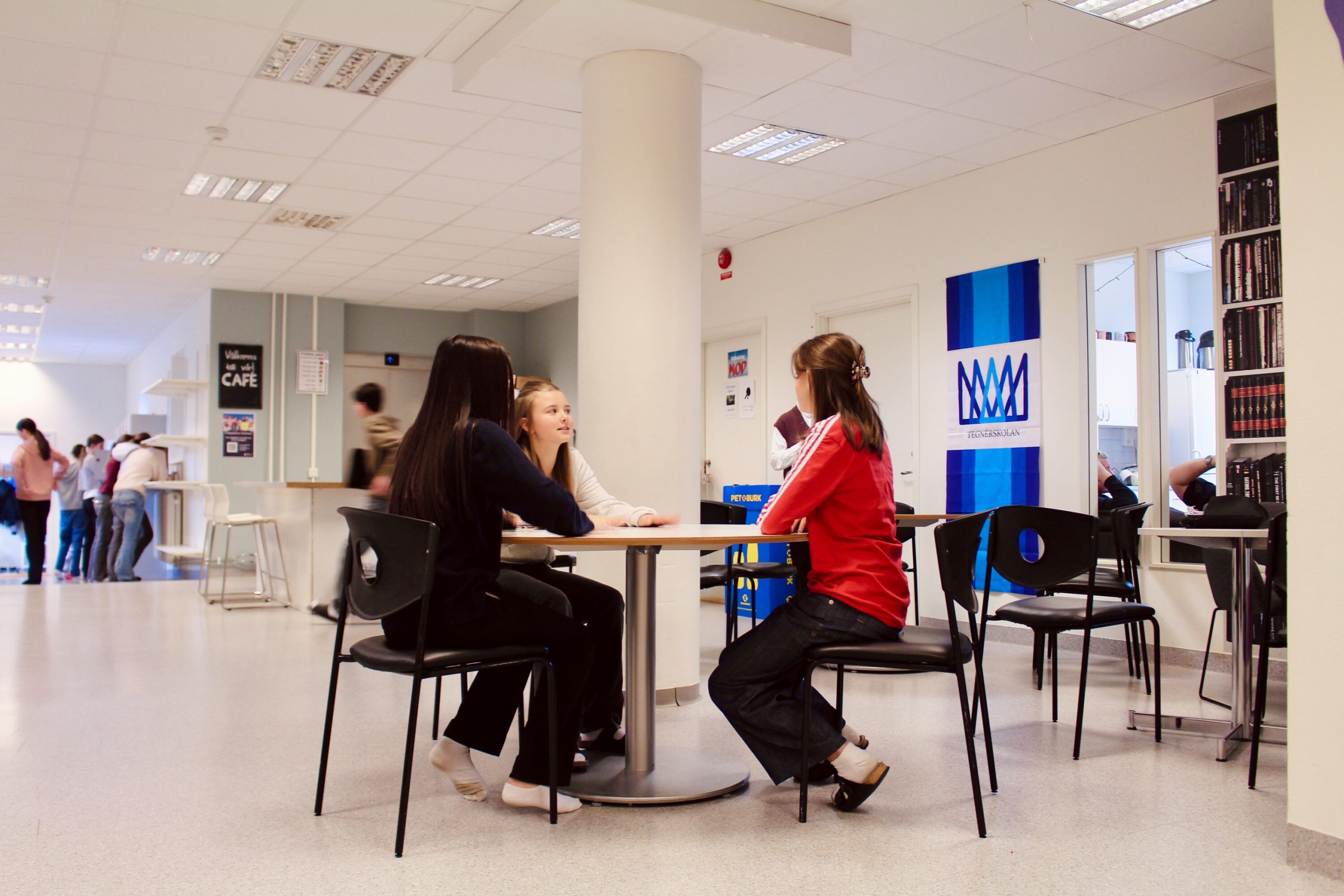 Elever sitter tillsammans och pratar vid ett runt bord i skolans gemensamma utrymme med kafé, en bokhylla och en banderoll med Tegnérskolans logotyp i bakgrunden.
