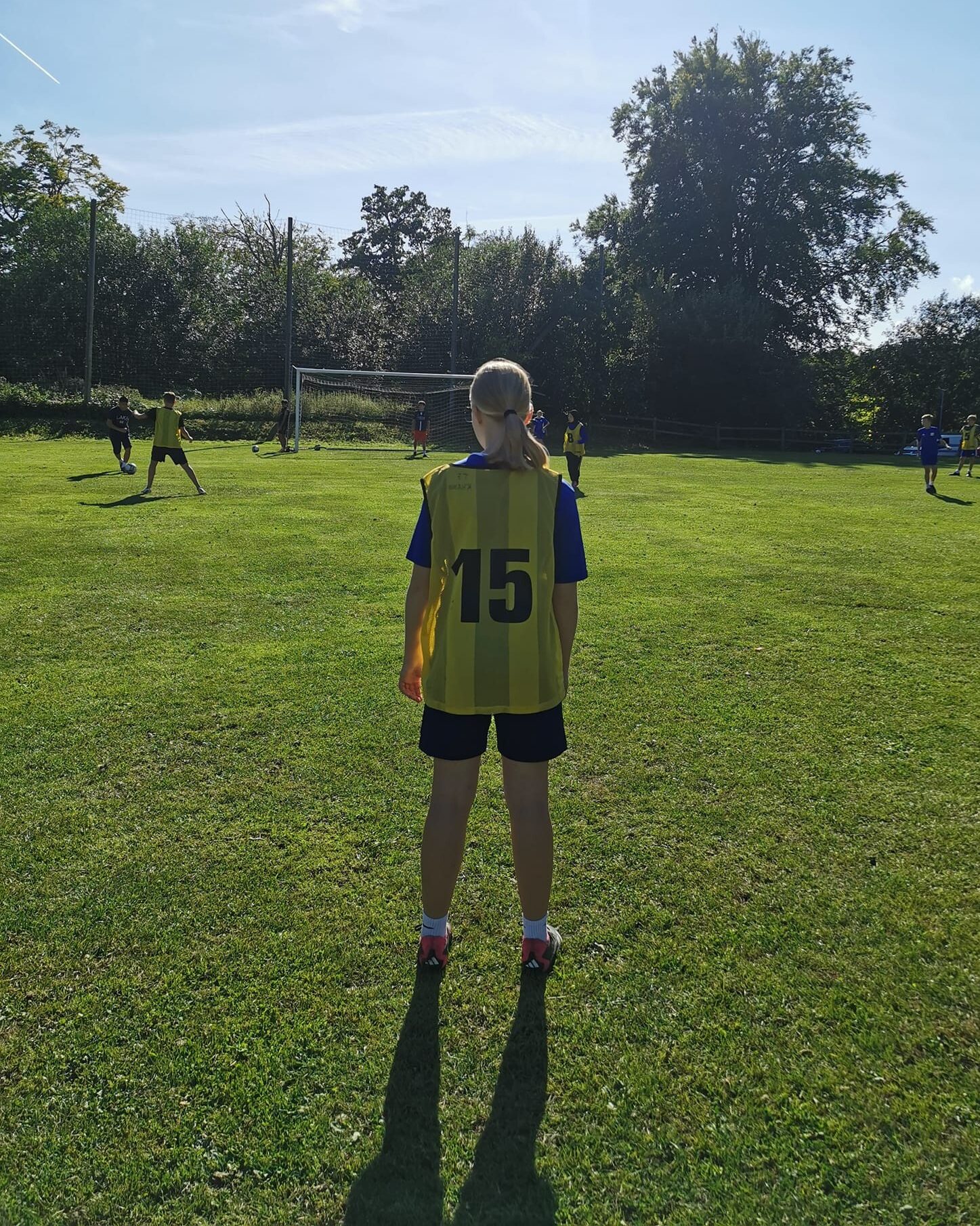 En spelare i gul västtröja med numret 15 står på en fotbollsplan och tittar mot spelet, med fler spelare synliga på planen en solig dag.