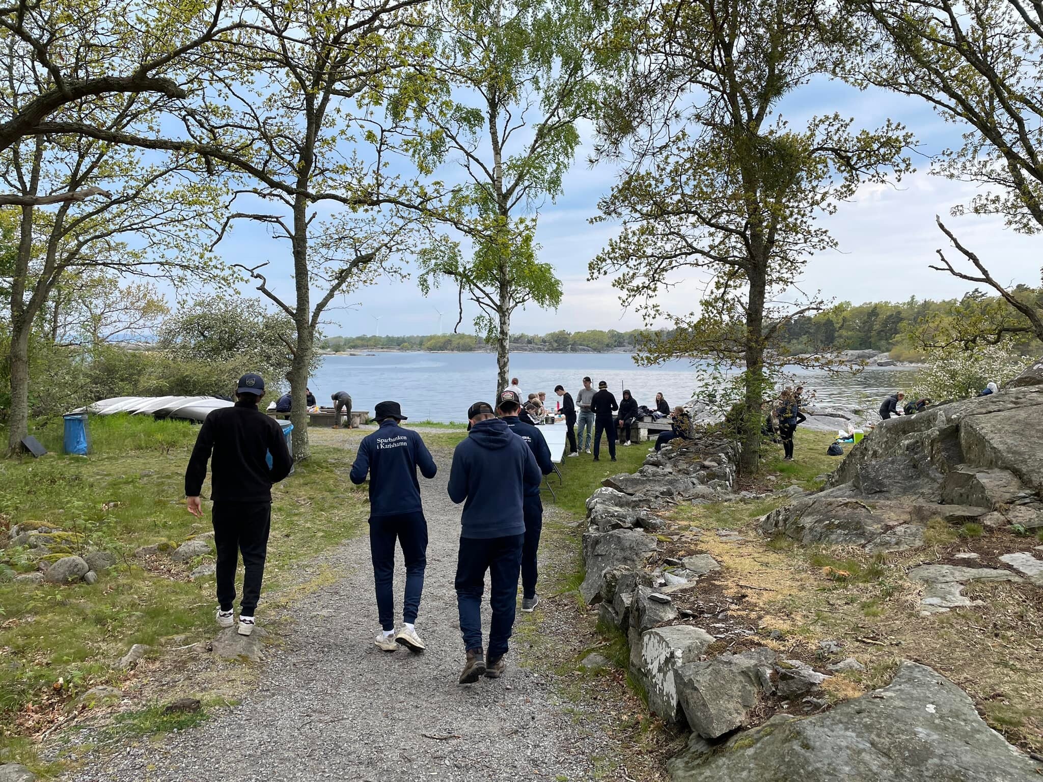Elever går tillsammans på en grusstig mot en samlingsplats vid havet, där fler personer syns vid bord och kanoter i ett klippigt kustlandskap.