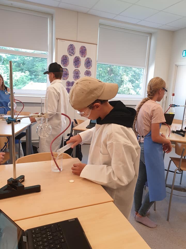 Elever i laboratorierockar utför ett kemiexperiment med glasflaskor och slangar i ett klassrum, med en elev i keps som skyddsglasögon hälls i en bägare.