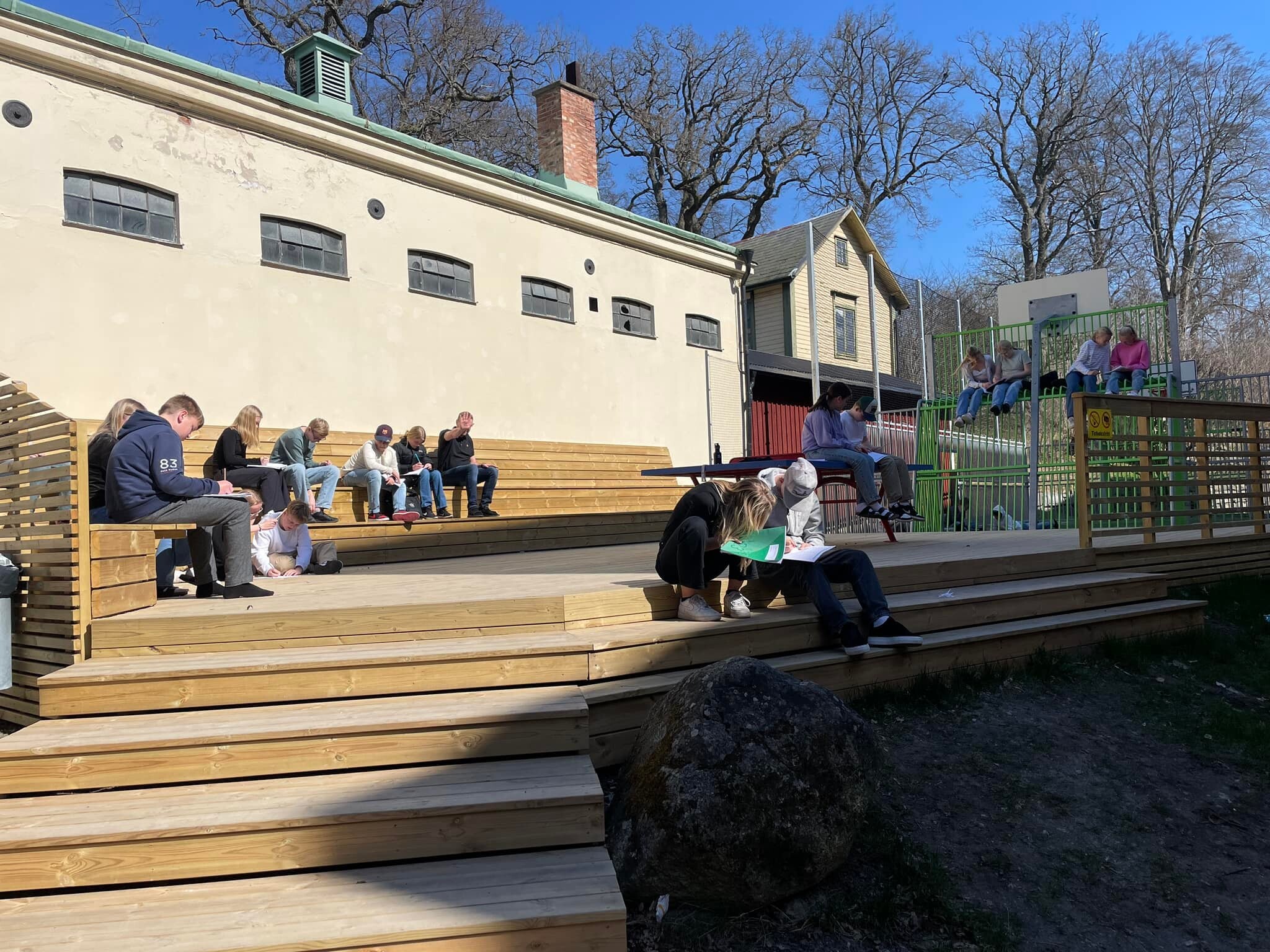 Elever sitter utomhus på en trätrappa och skriver i sina anteckningsböcker under en utomhuslektion en solig dag, med en gammal byggnad och kala träd i bakgrunden.